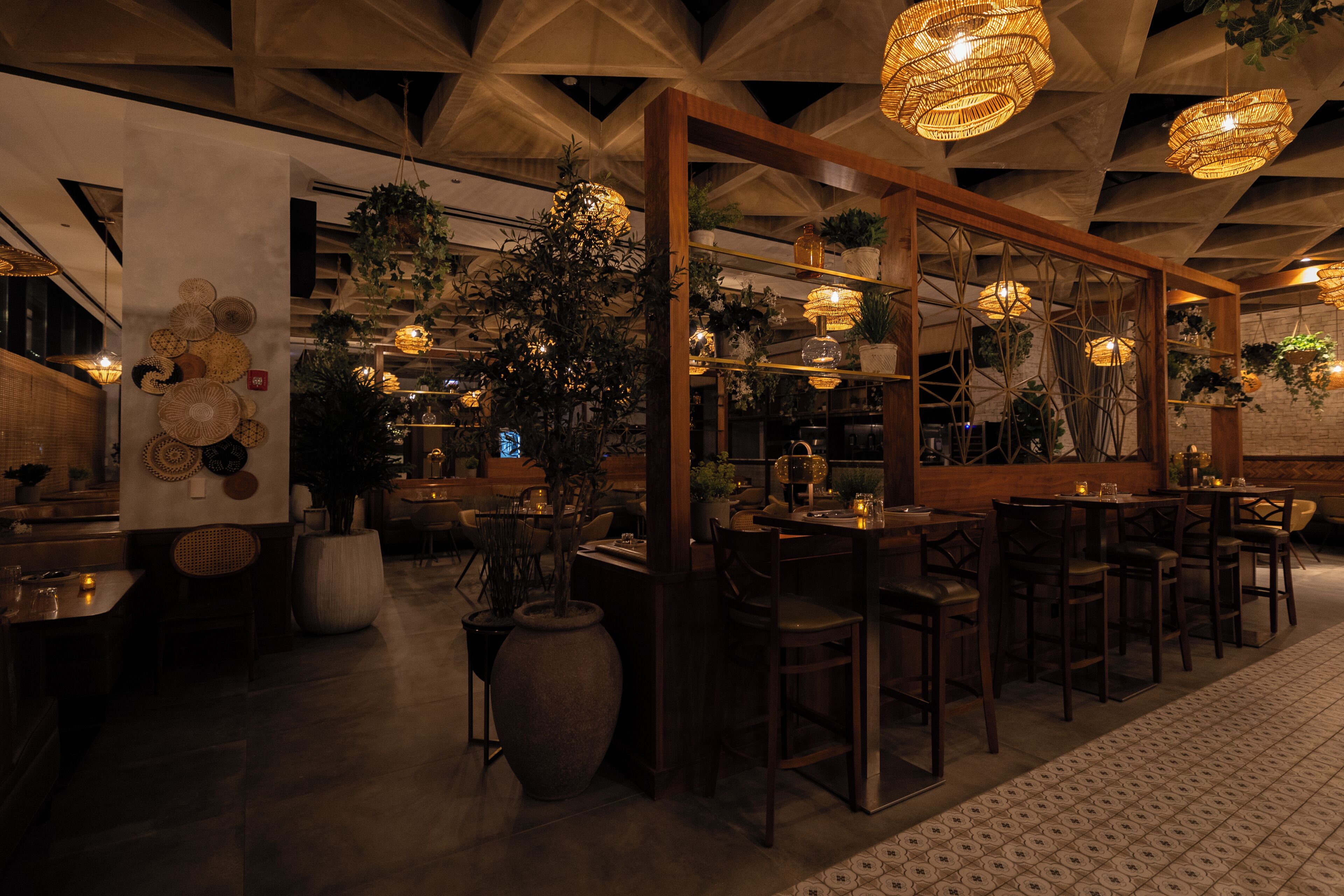 Interior de restaurante com mesas e cadeiras de madeira, plantas pendentes e padrões geométricos no teto.
