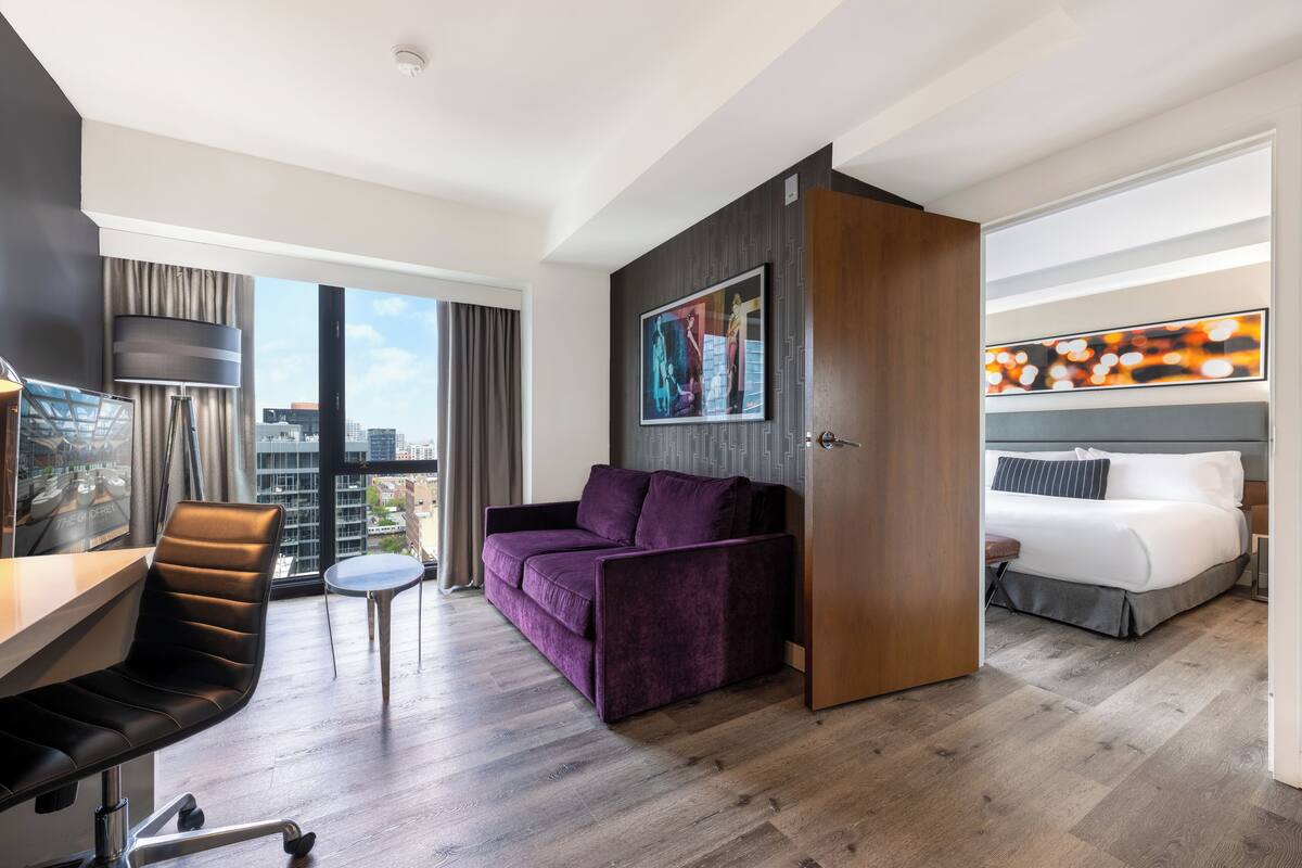 A modern hotel room with a large bed, a purple sofa, a desk with a chair, and a city view.
