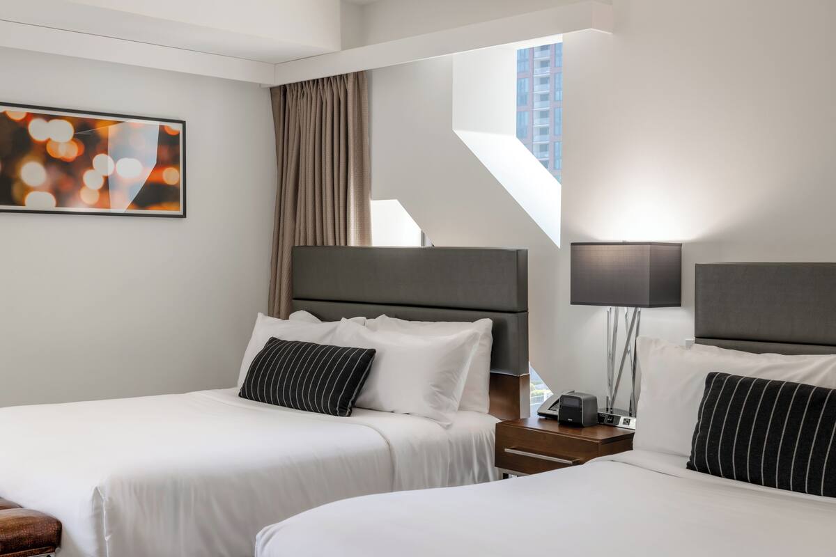 A modern hotel room with two beds, a large abstract painting, a view of the city, and a staircase.
