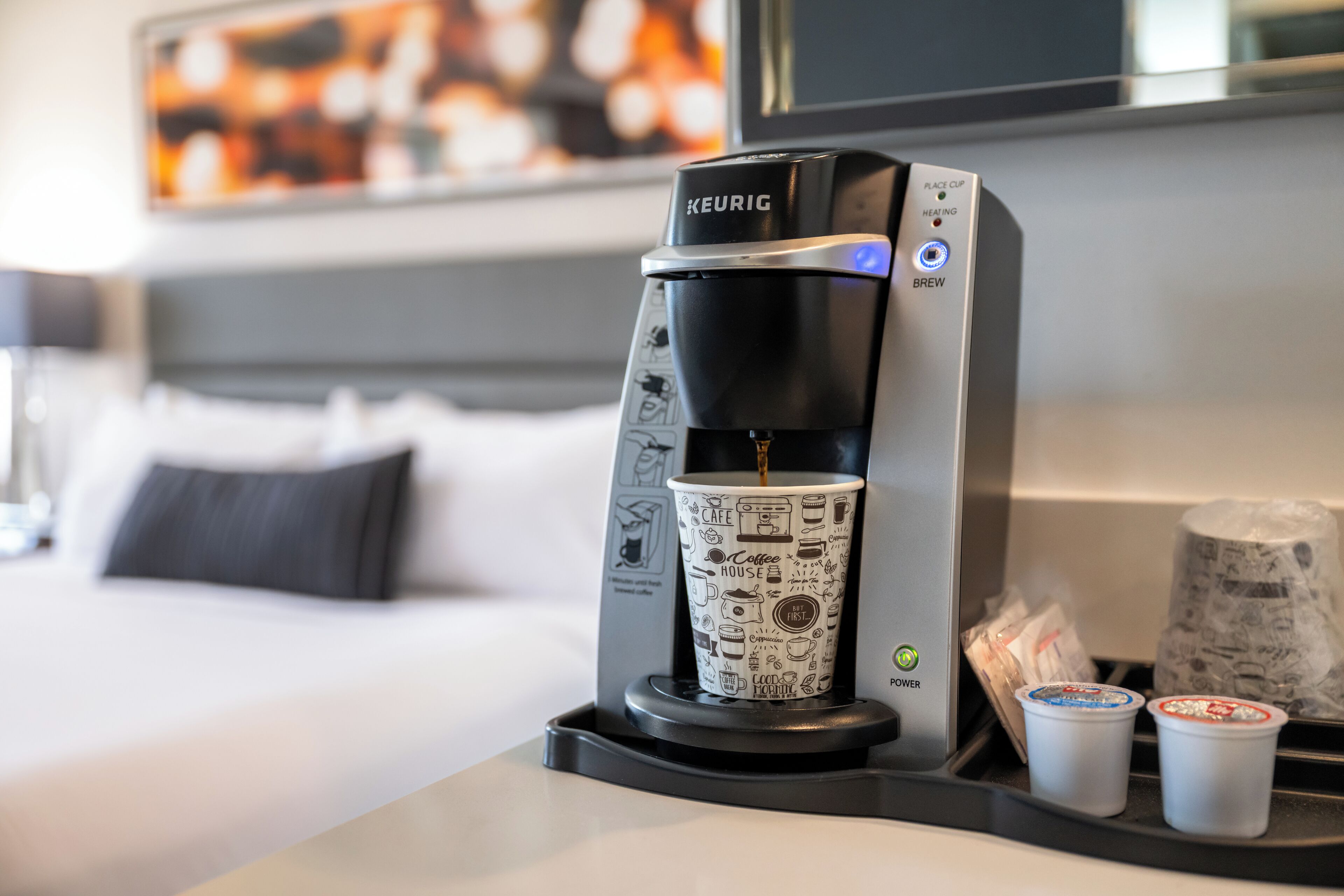 Une machine à café Keurig avec un porte-tasse, deux gobelets jetables et un lit en arrière-plan.