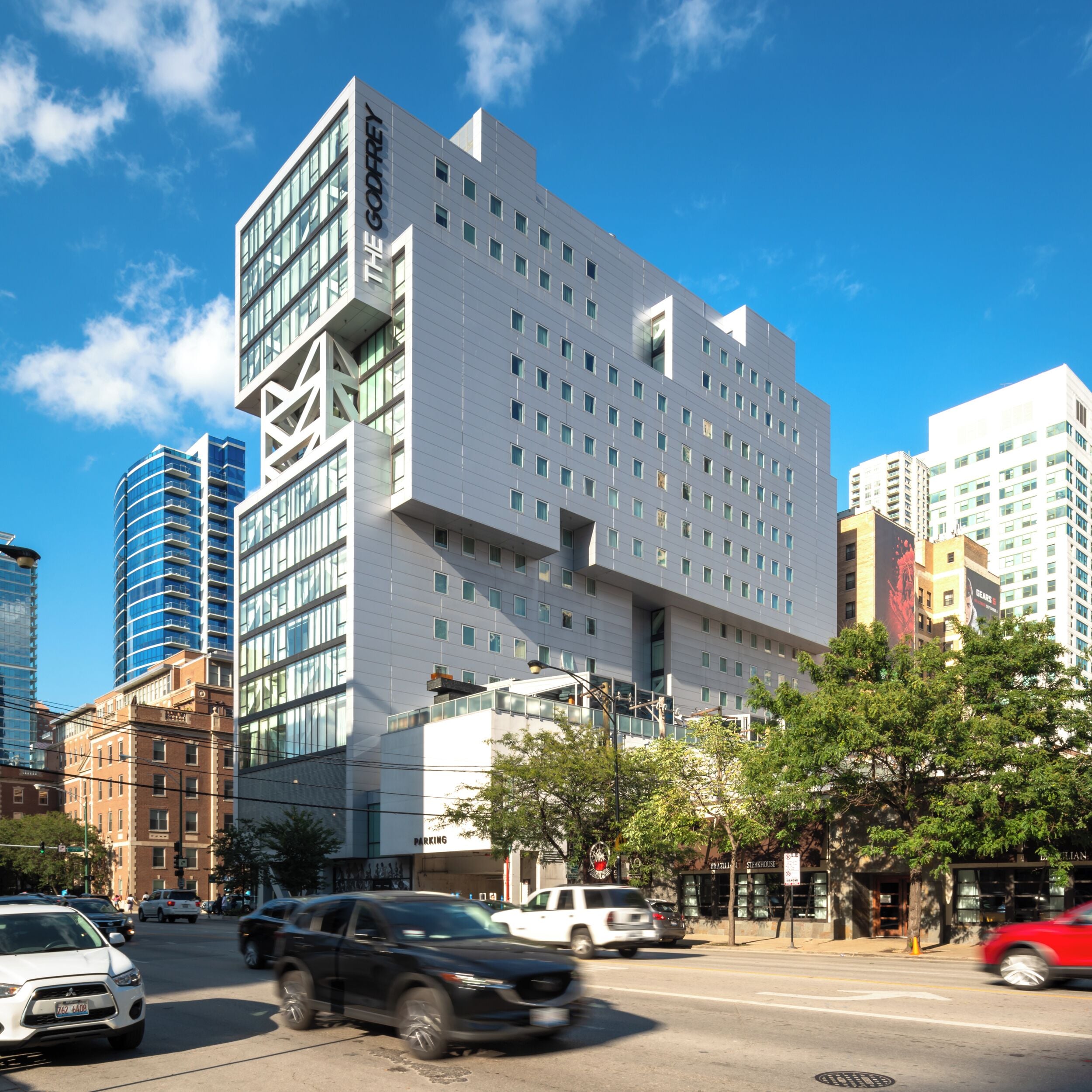 Un edificio moderno con un llamativo letrero 'GODFREY', rodeado de otros rascacielos y una calle concurrida llena de coches.