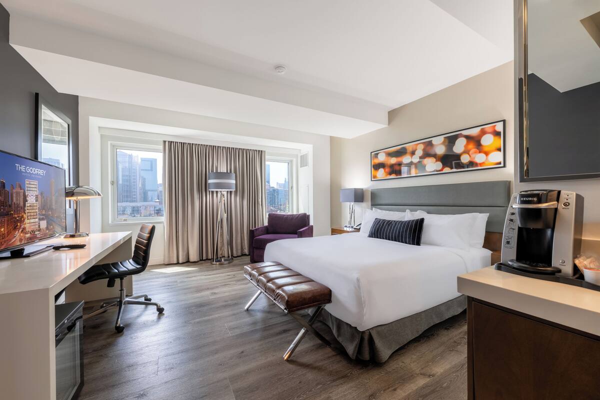 A hotel room with a large bed, a desk with a computer, a chair, a bench, a coffee maker, and a city view.