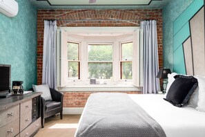 A bedroom with a brick wall, a bay window, a bed, a desk, a chair, and a TV.