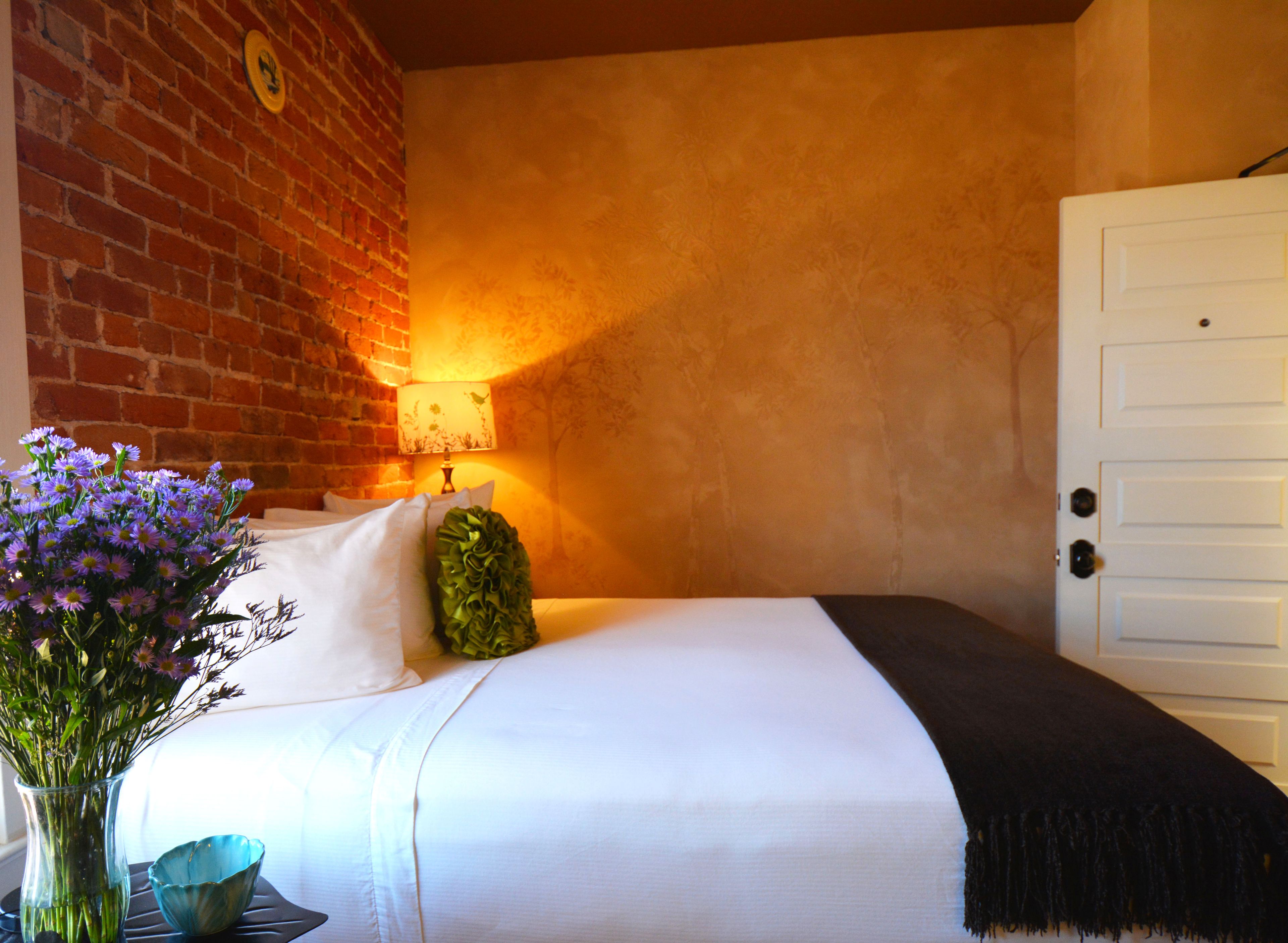 Quarto de hotel com parede de tijolos, cama com roupa de cama branca, mesa de cabeceira com abajur e um vaso de flores.