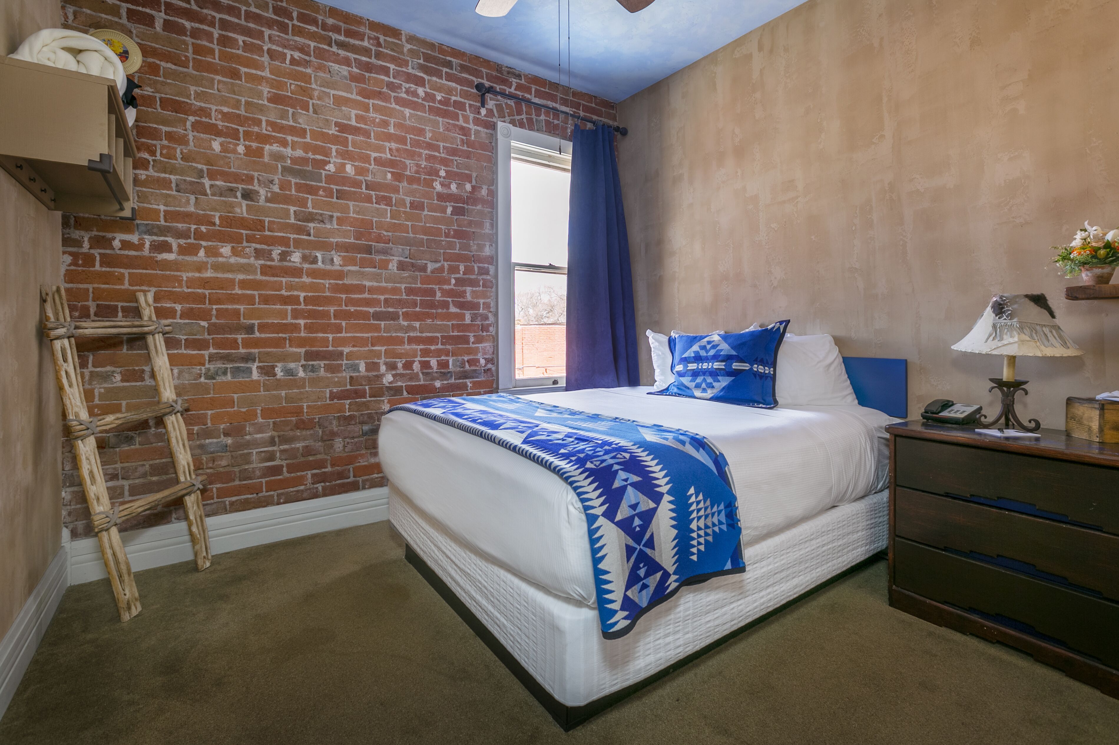 A bedroom with a brick wall, a bed with a blue patterned blanket, a wooden ladder, a dresser, and a window with blue curtains.