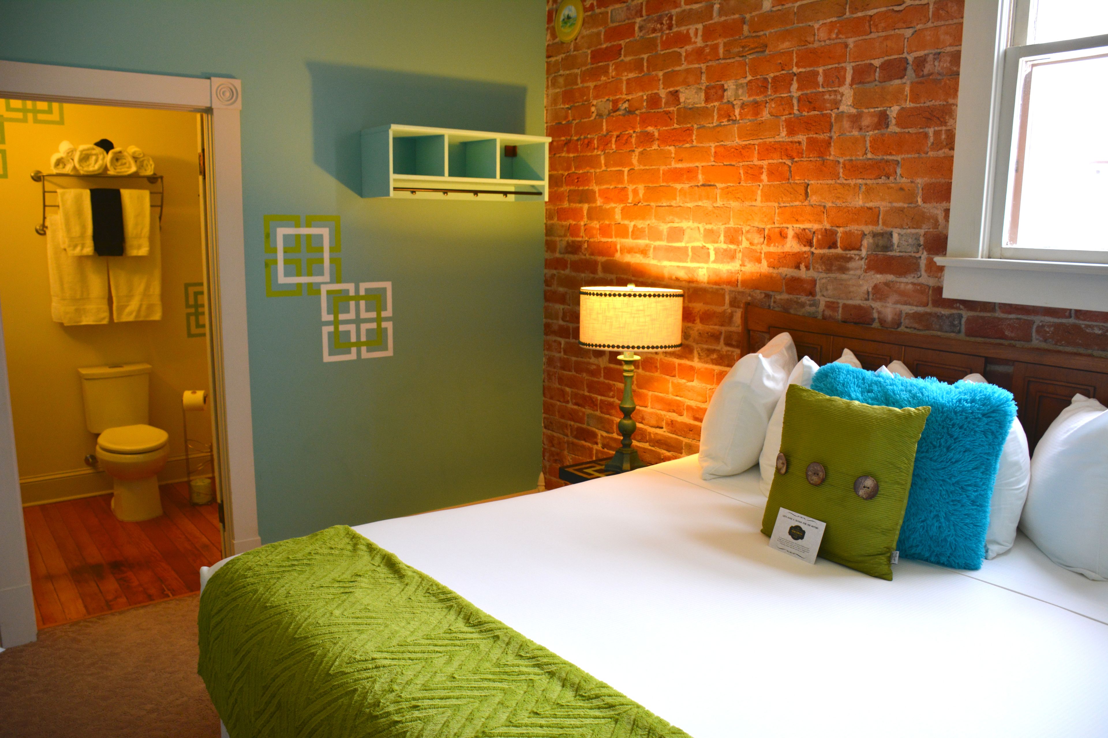Une chambre d’hôtel avec une couvre-lit à motifs chevrons verts, un mur en briques, une tête de lit en bois et une table de chevet avec une lampe.