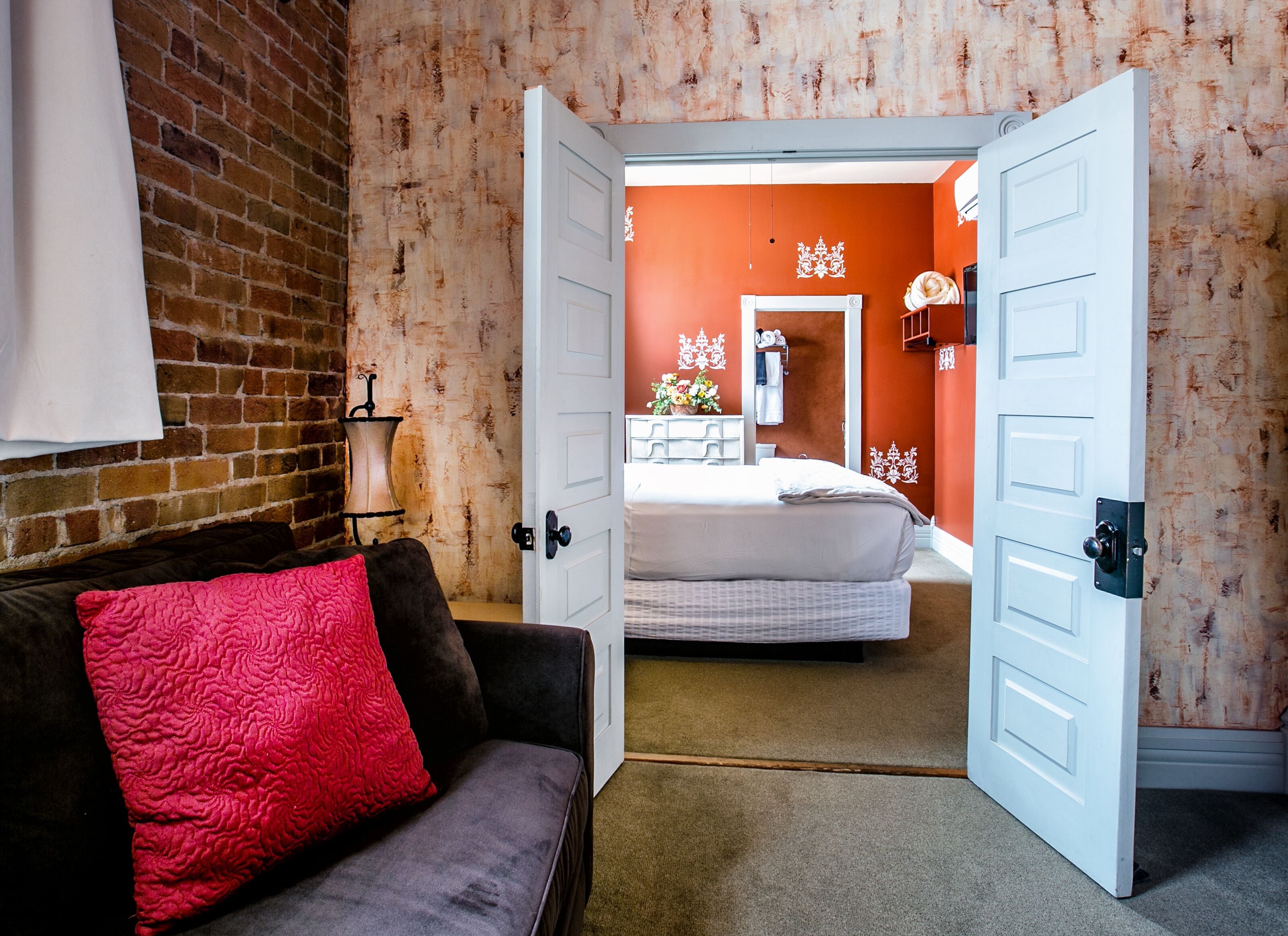 Una habitación con pared de ladrillo, una puerta blanca que da a un dormitorio, un cojín rojo sobre un sofá oscuro y una lámpara blanca.