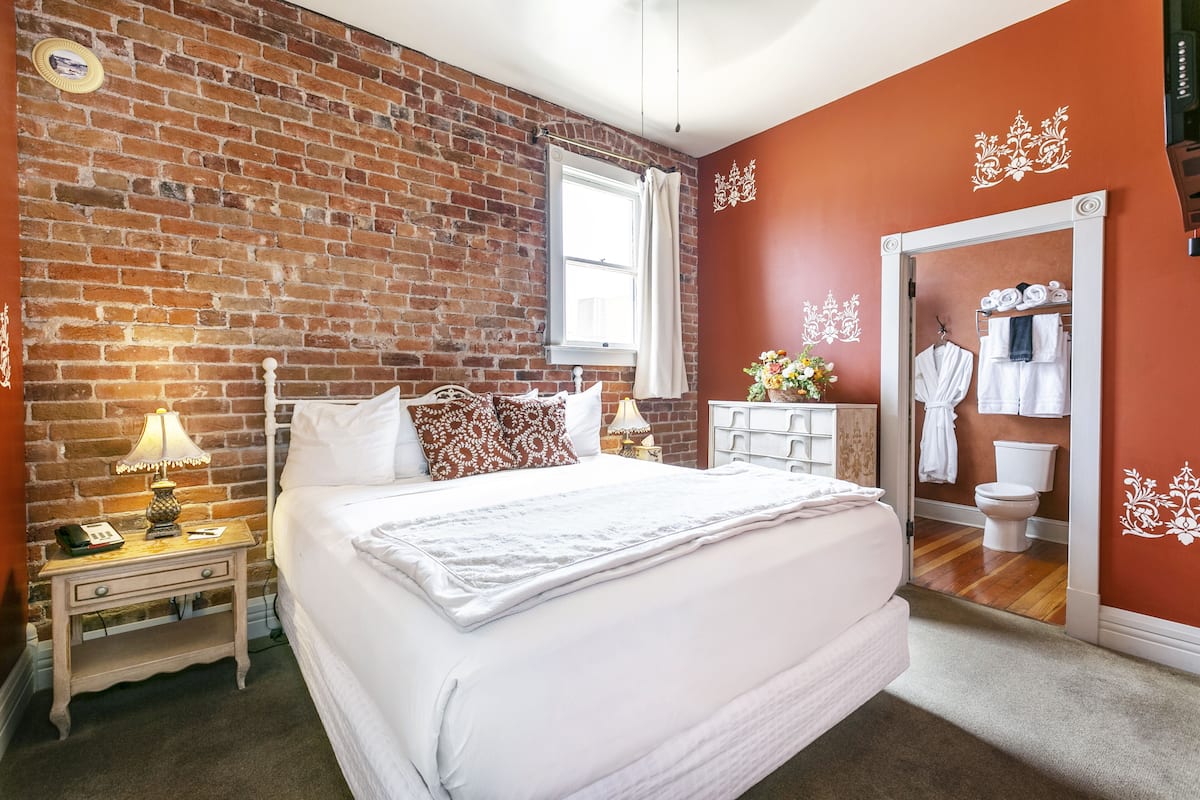 A bedroom with a brick wall, a large bed, a nightstand with a lamp, a dresser, and a mirror.