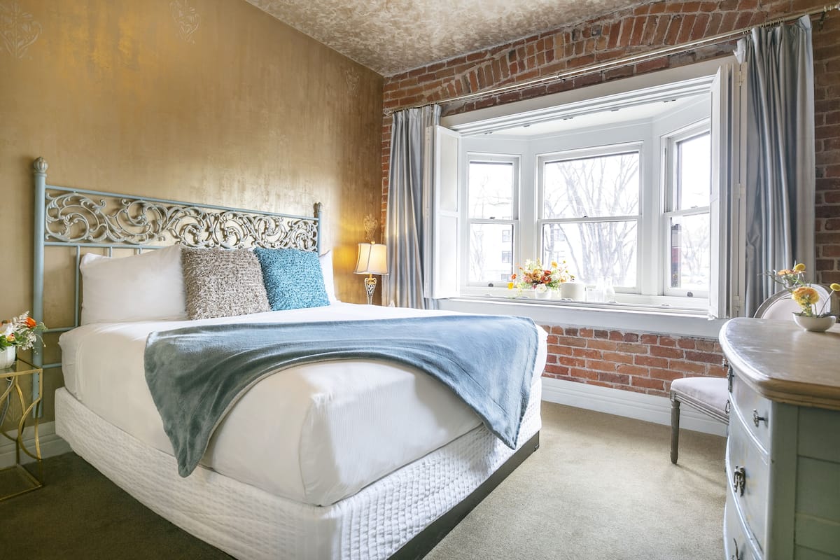 A bedroom with a bed, a nightstand, a dresser, a window with curtains, and a brick wall.