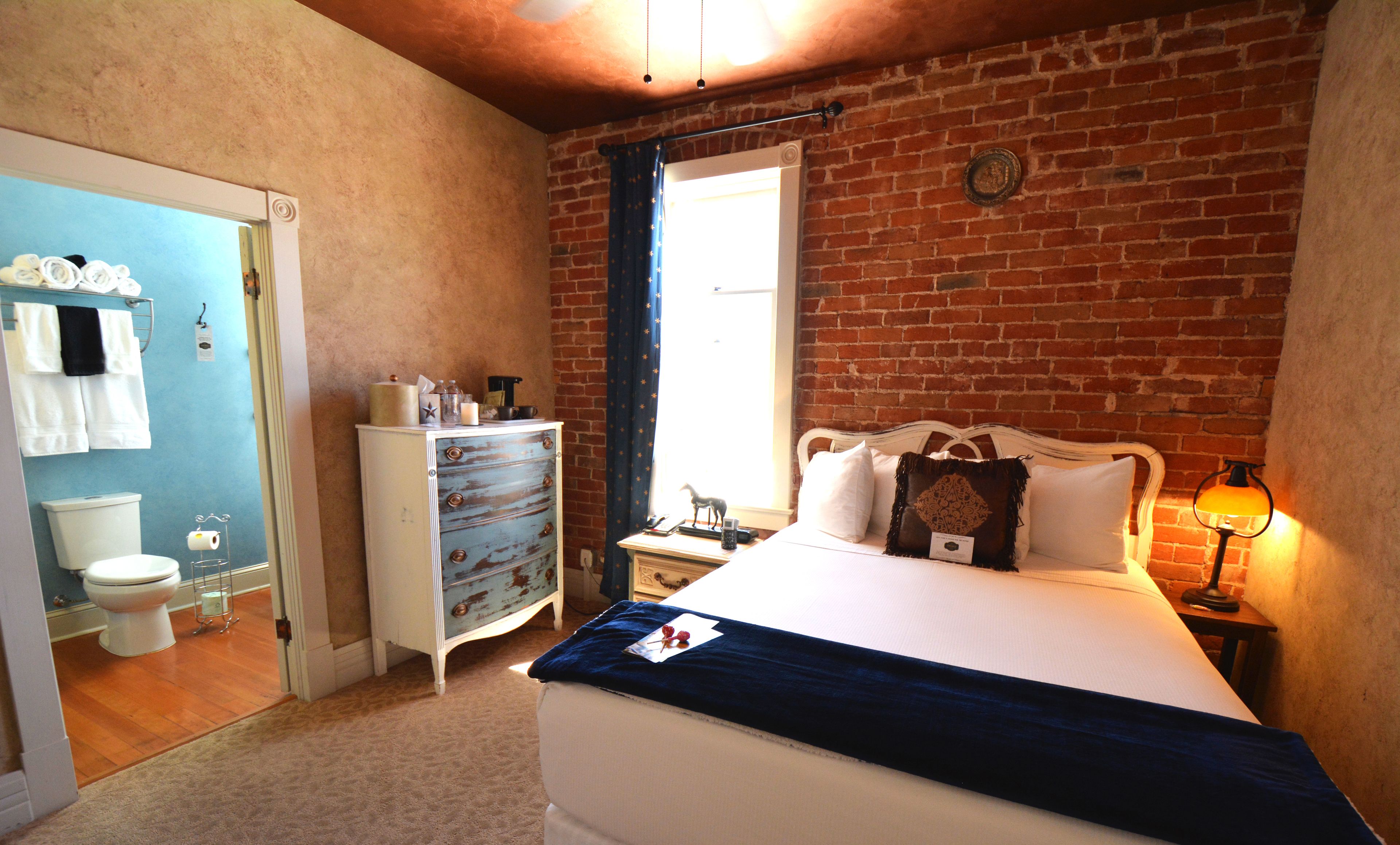 A bedroom with a brick wall, a bed with white bedding, a nightstand with a lamp, a dresser, and a window with curtains.