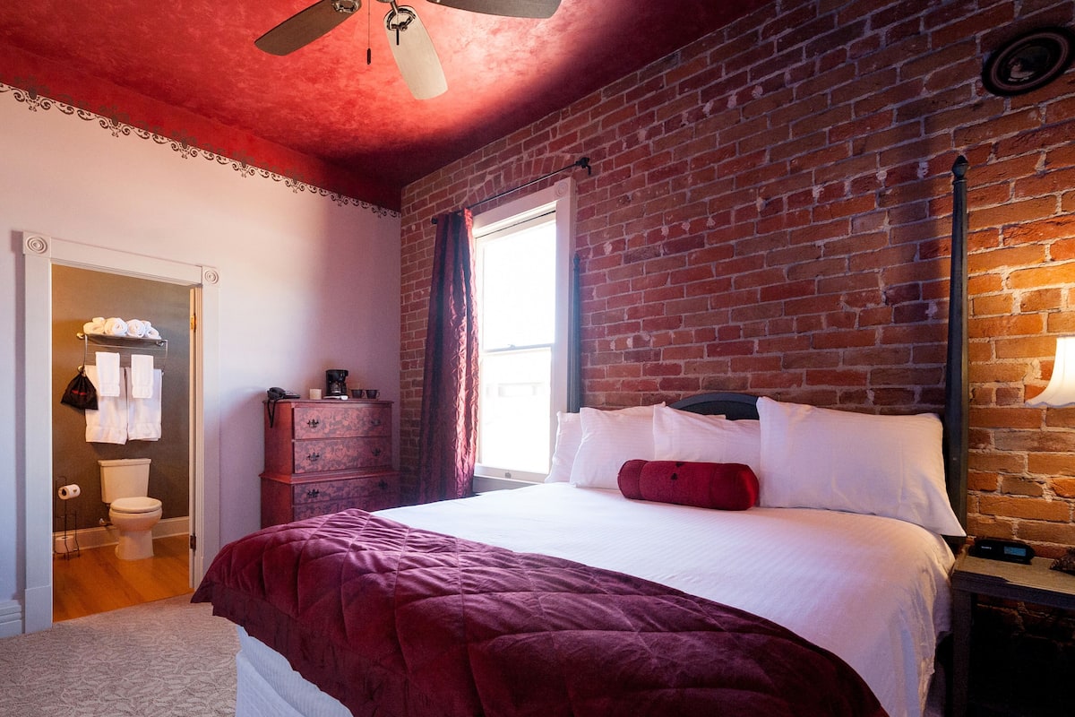 A bedroom with a brick wall, a bed with white bedding, a red throw, a dresser, and a window with curtains.
