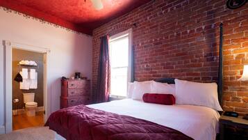 A bedroom with a brick wall, a bed with white bedding, a red throw, a dresser, and a window with curtains.
