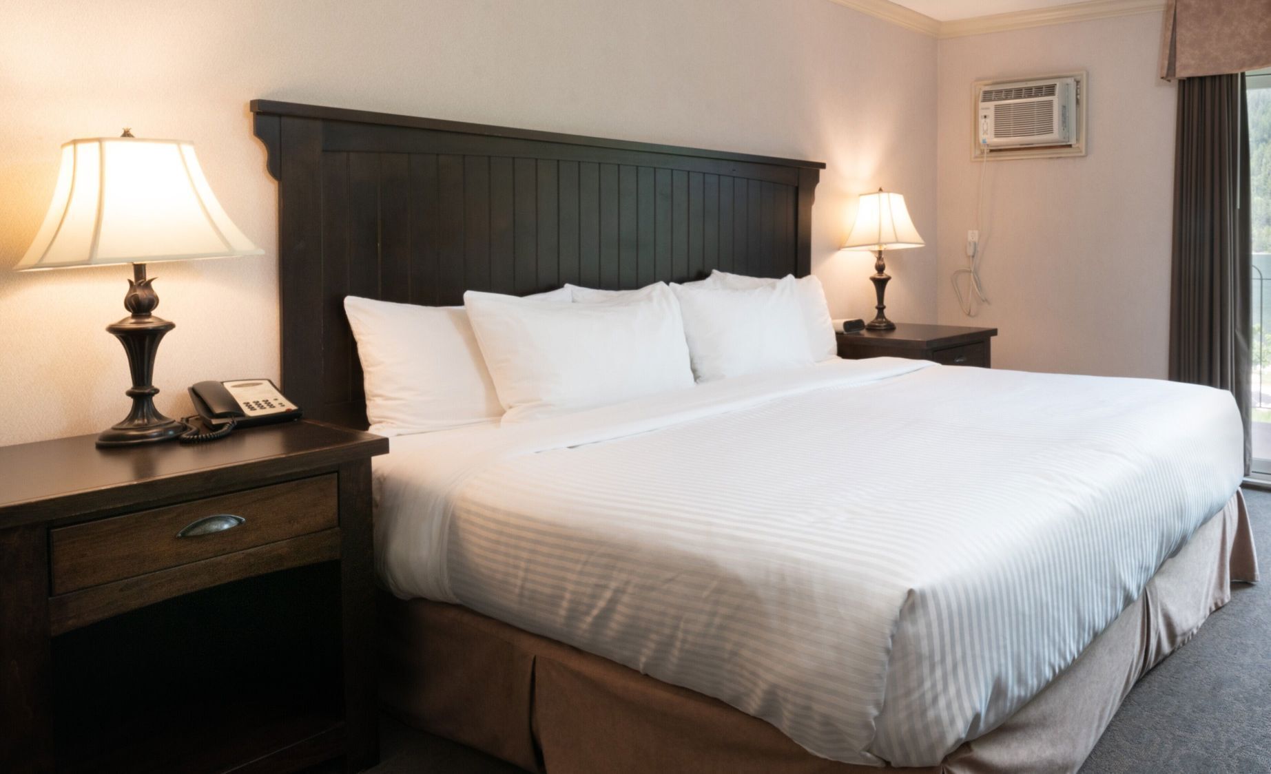A hotel room with a large bed, two bedside tables with lamps, a telephone, and an air conditioning unit.