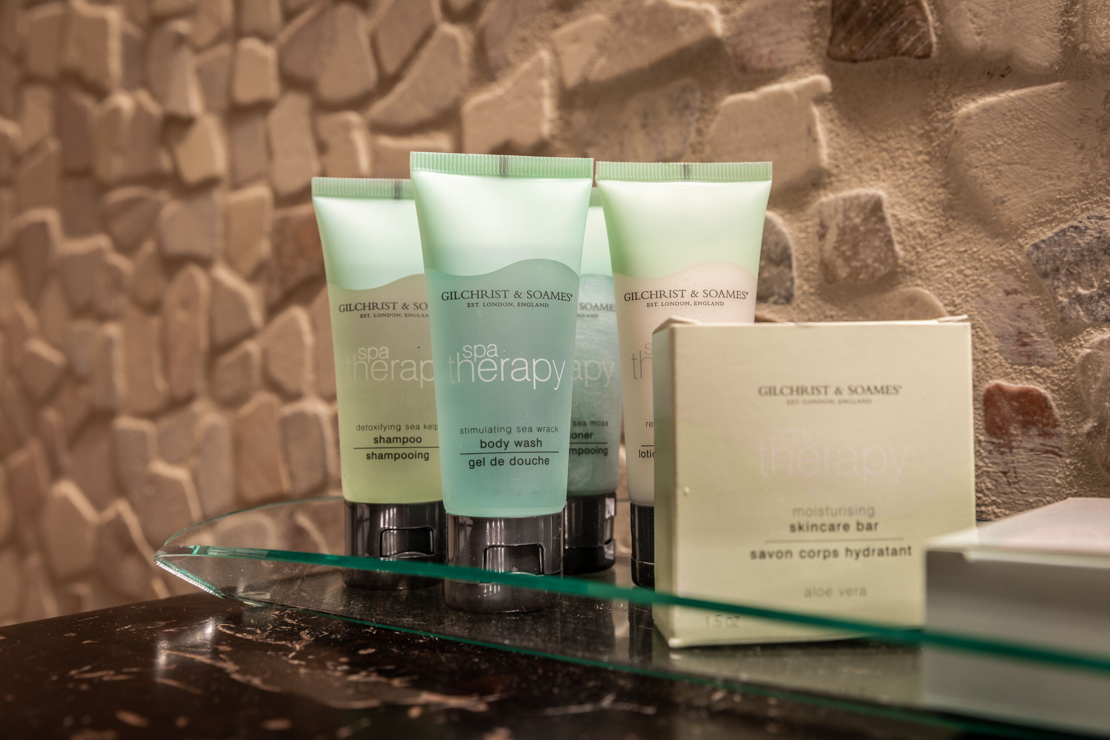 A display of Gilchrist & Soames spa therapy products on a glass shelf against a textured wall.
