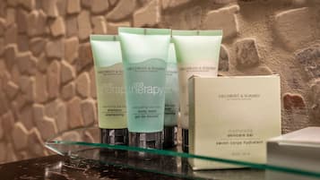 Une sélection de produits de soins de spa de la marque Gilchrist & Soames est exposée sur une étagère en verre, le tout mis en valeur par un mur à texture.