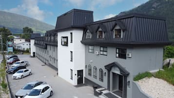 A hotel with a white facade and dark roof, a parking lot with several cars, and a mountainous backdrop.