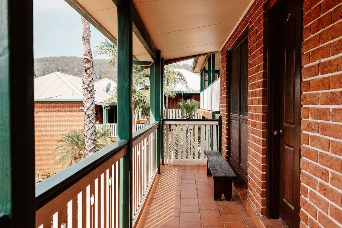 A covered balcony with a bench, brick walls, and a view of neighboring buildings and palm trees.