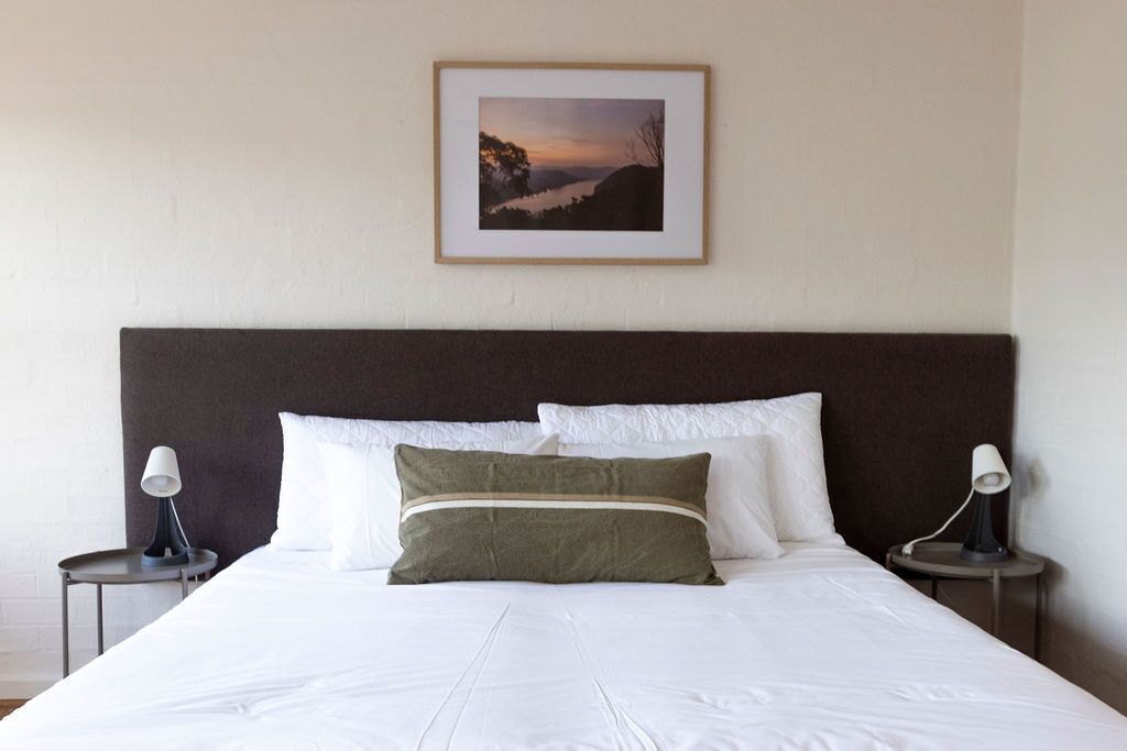 A neatly made bed with white linens and two pillows, a framed picture on the wall, and two bedside lamps on either side.