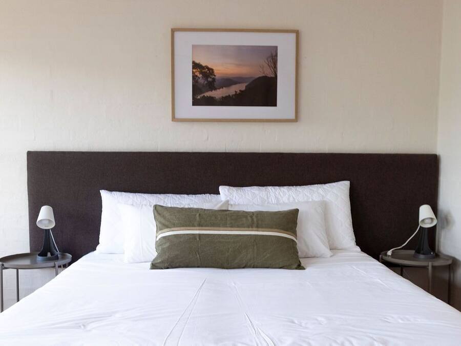 A neatly made bed with white linens and two pillows, a framed picture on the wall, and two bedside lamps on either side.