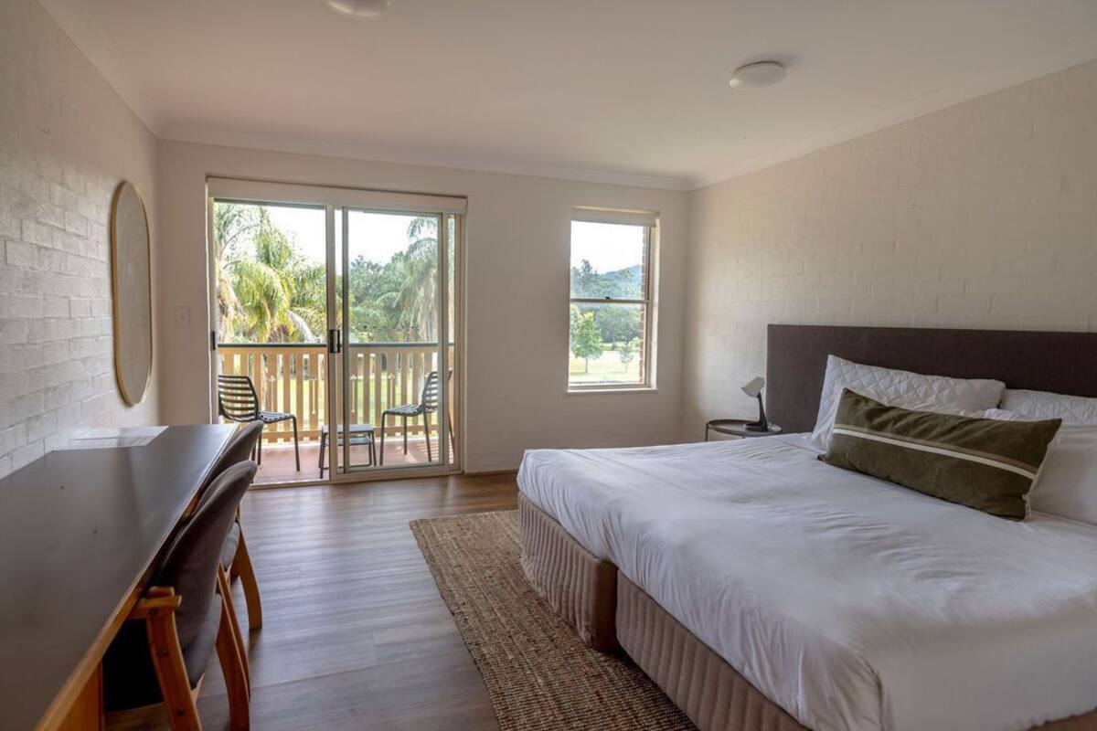 A bedroom with a large bed, a desk, and a balcony with chairs.