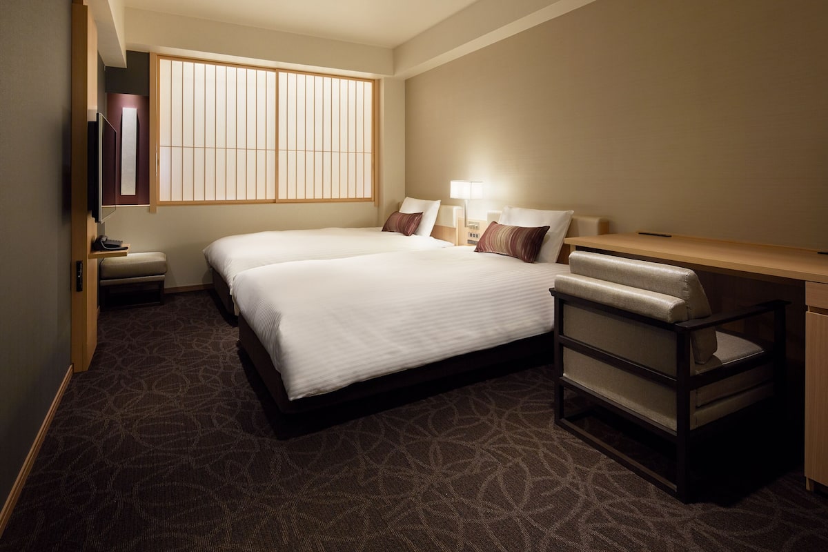 Ein Hotelzimmer mit einem großen Bett, einem Schreibtisch, einem Stuhl, einem Fernseher und einem Fenster mit vertikalen Jalousien.