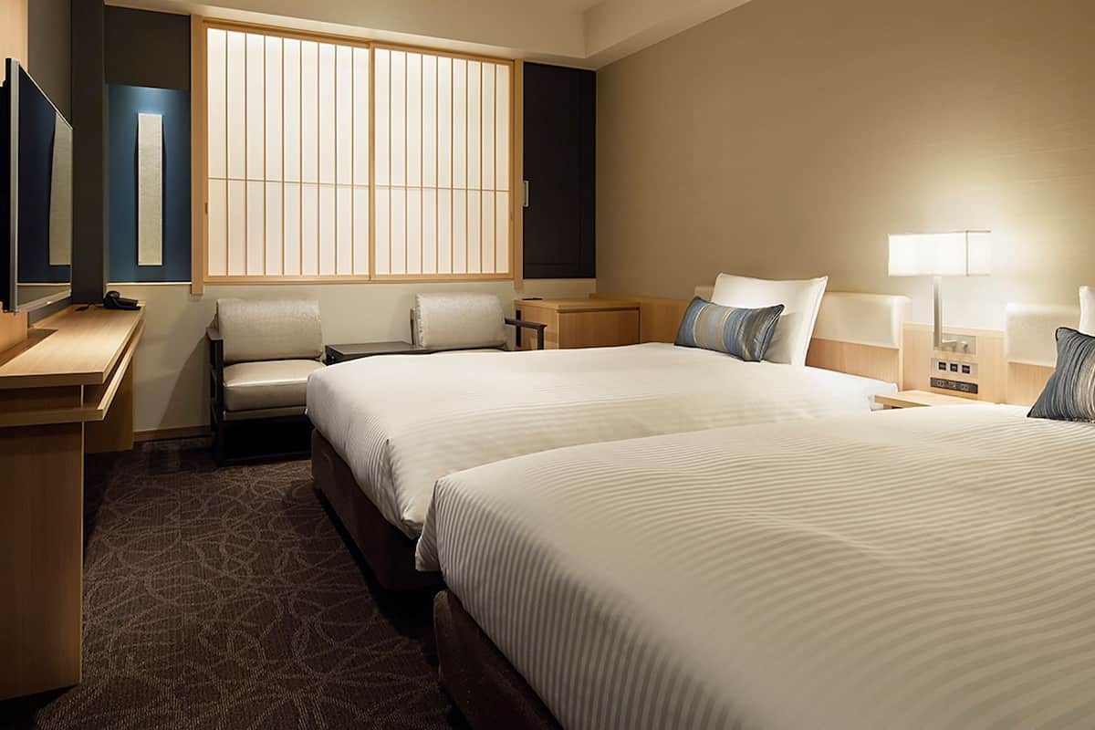 Ein Hotelzimmer mit zwei Betten, einem Schreibtisch, einem Sessel, einem Fernseher und einem Fenster mit Jalousien.
