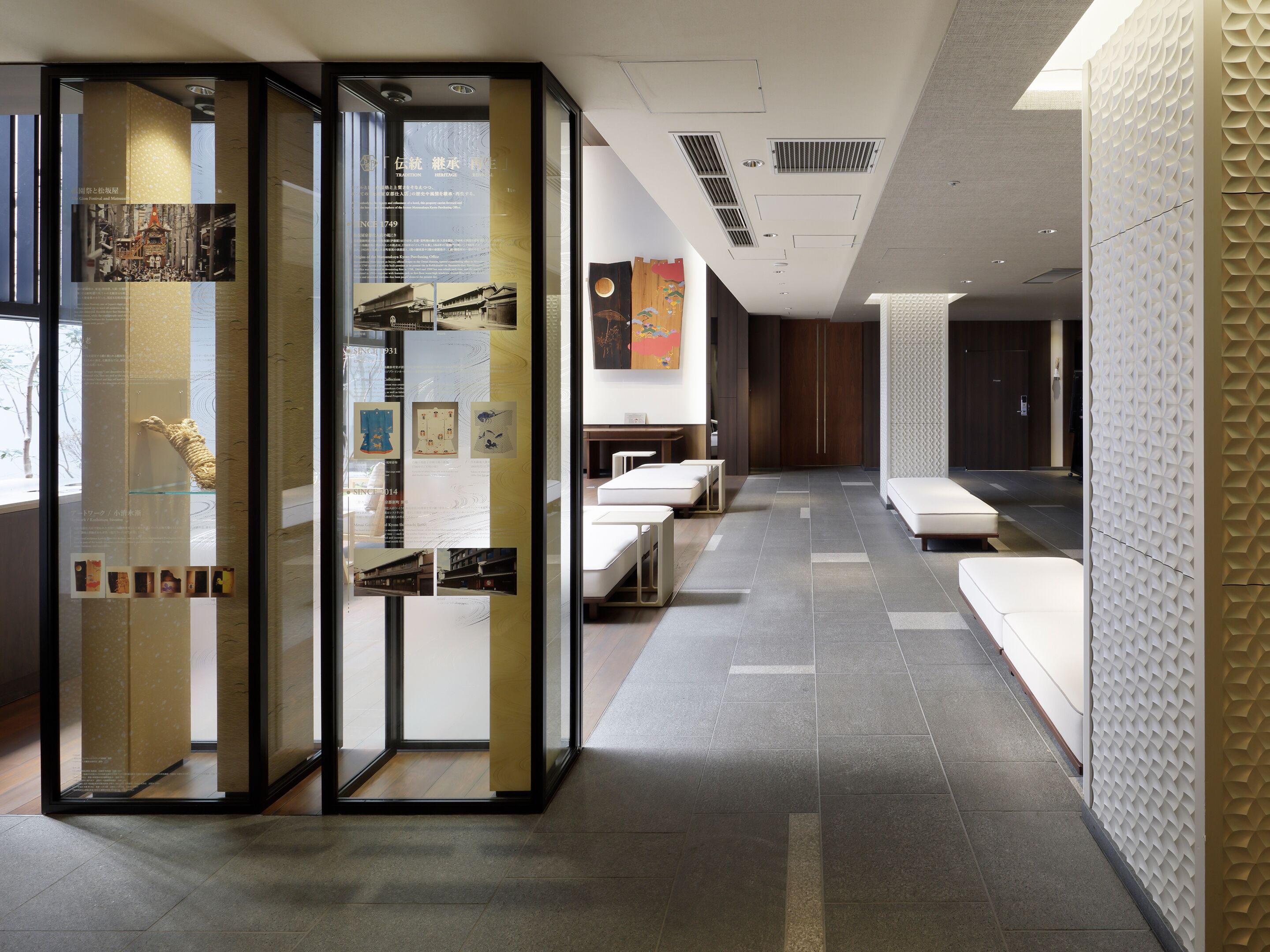 A modern hotel lobby with a reception desk, seating area, and a glass partition with informational displays.