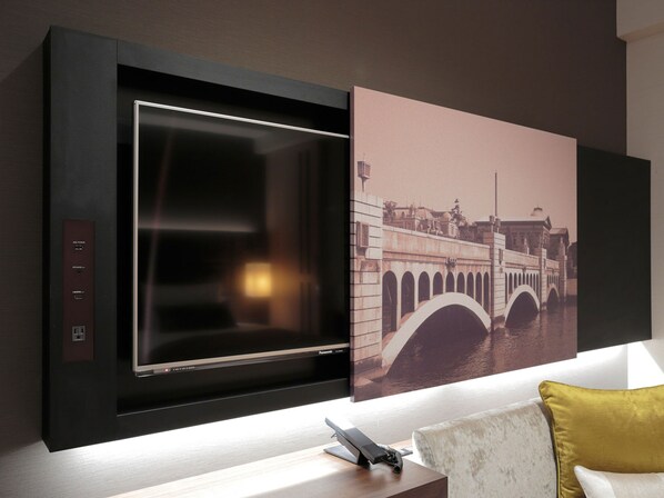 Un salón moderno con un televisor de pantalla plana grande montado en una pared oscura, un cuadro de un puente y un escritorio pequeño con un teléfono.