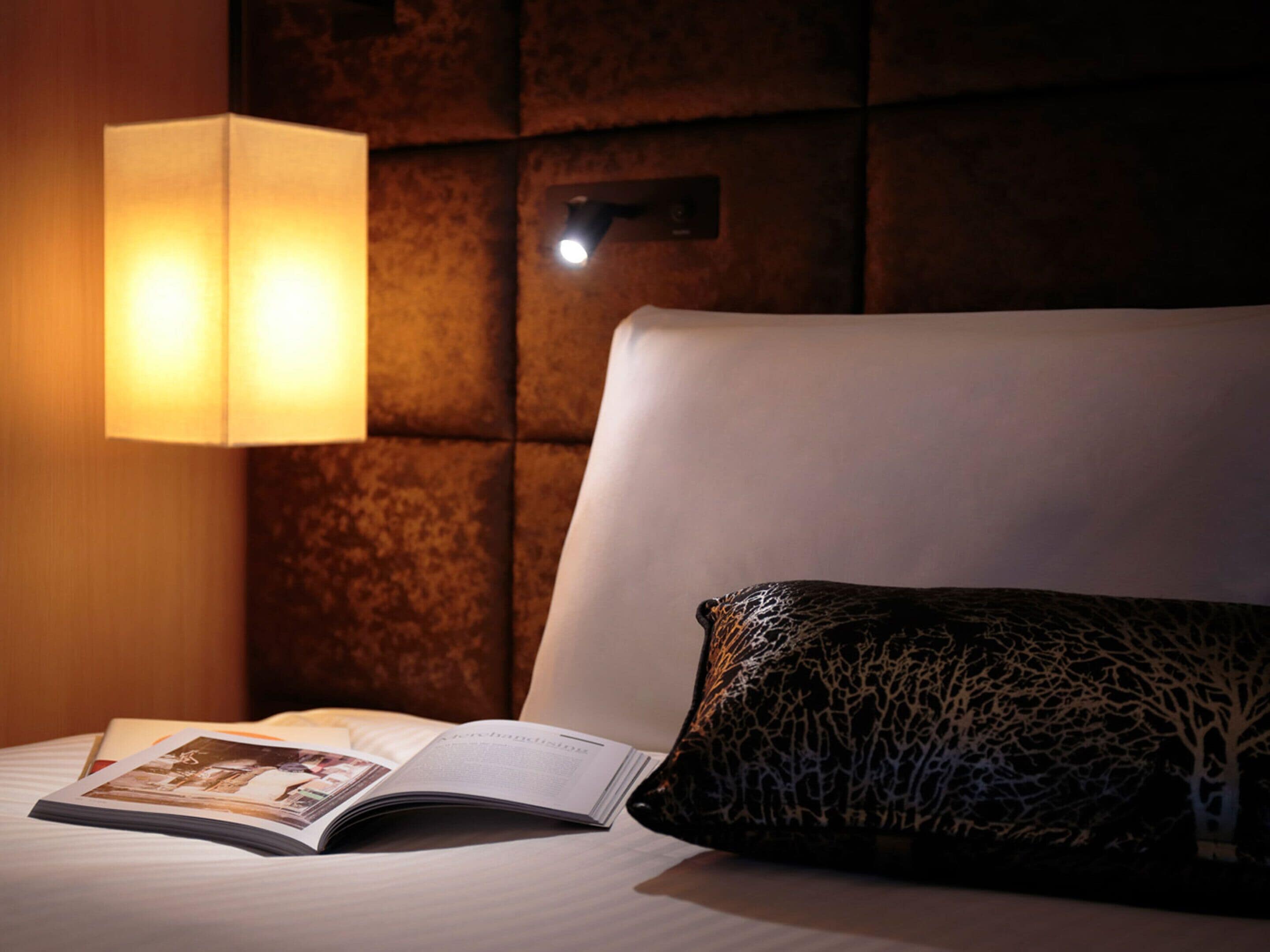 A hotel room with a bed, a book, a patterned pillow, a wall-mounted lamp, and a textured wall.