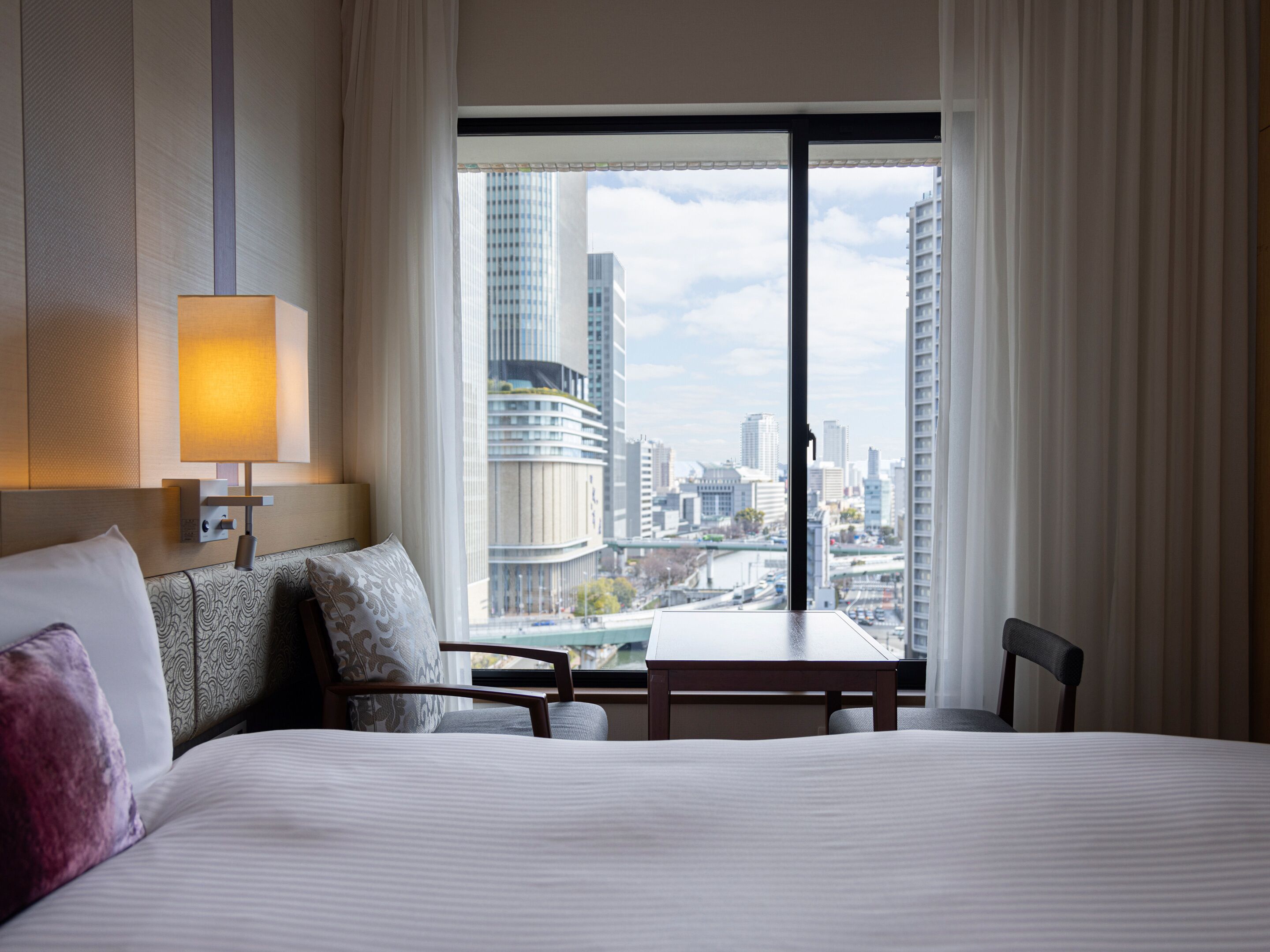 A hotel room with a large bed, a bedside table, a chair, a lamp, and a view of the cityscape through a large window.