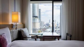 A hotel room with a large bed, a bedside table, a chair, a lamp, and a view of the cityscape through a large window.