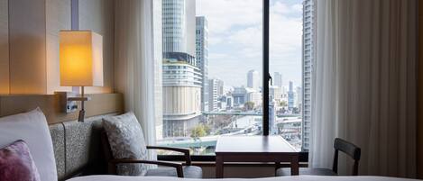 A hotel room with a large bed, a bedside table, a chair, a lamp, and a view of the cityscape through a large window.