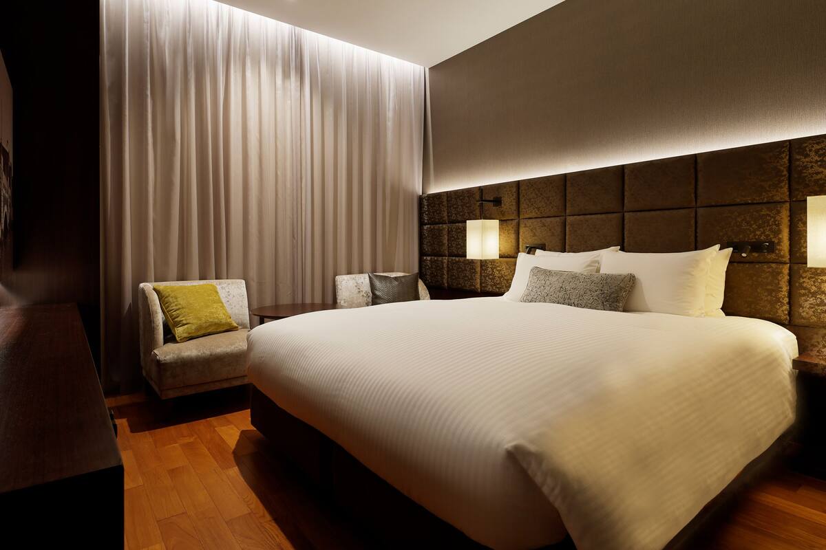 Une chambre d’hôtel avec un grand lit, un petit fauteuil, une table de chevet et une lampe fixée au mur.