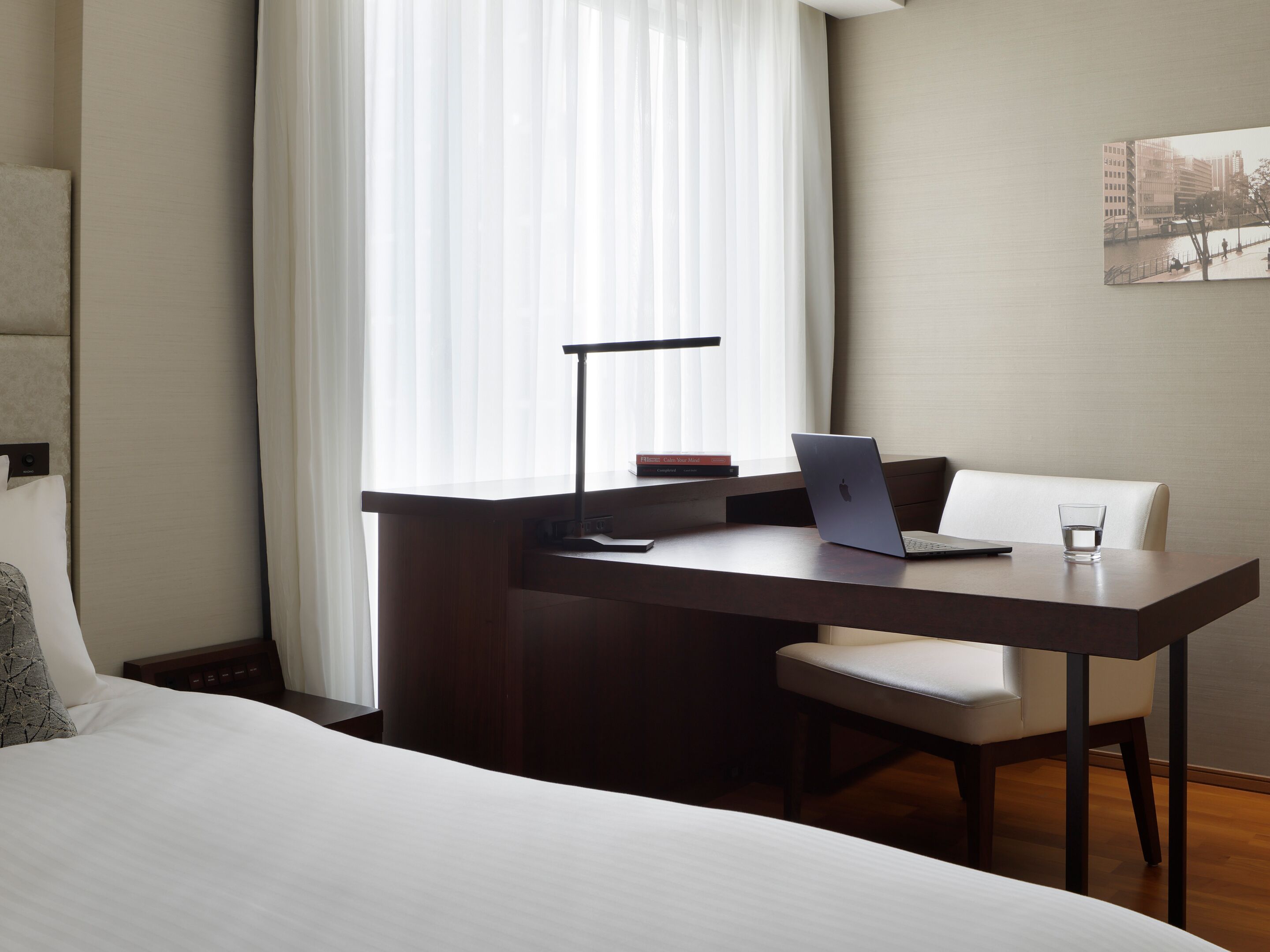 A hotel room with a bed, a desk with a laptop, a chair, a lamp, and a glass of water.