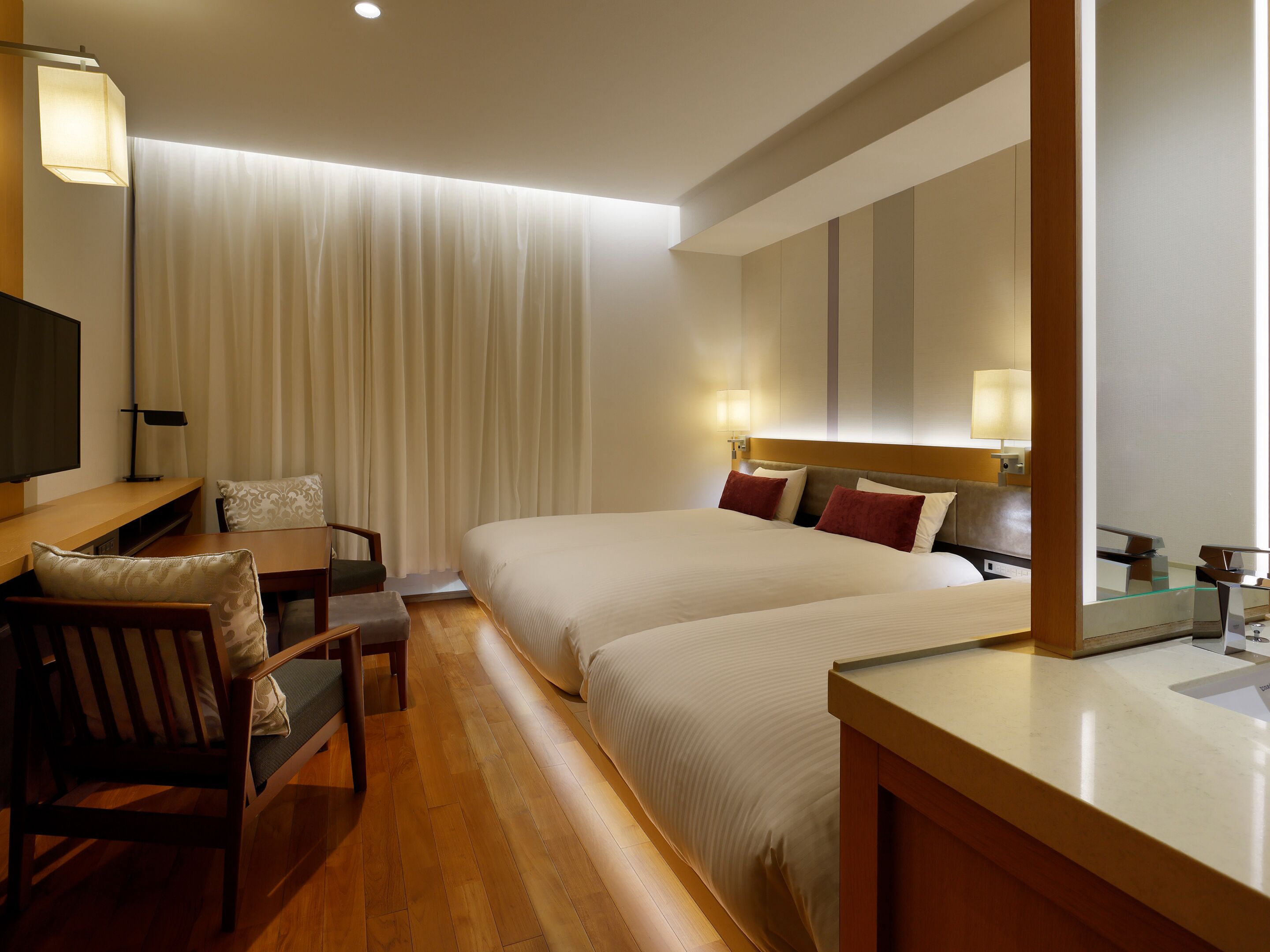 A hotel room with a bed, bedside tables, a desk with a chair, a TV, and a window with curtains.