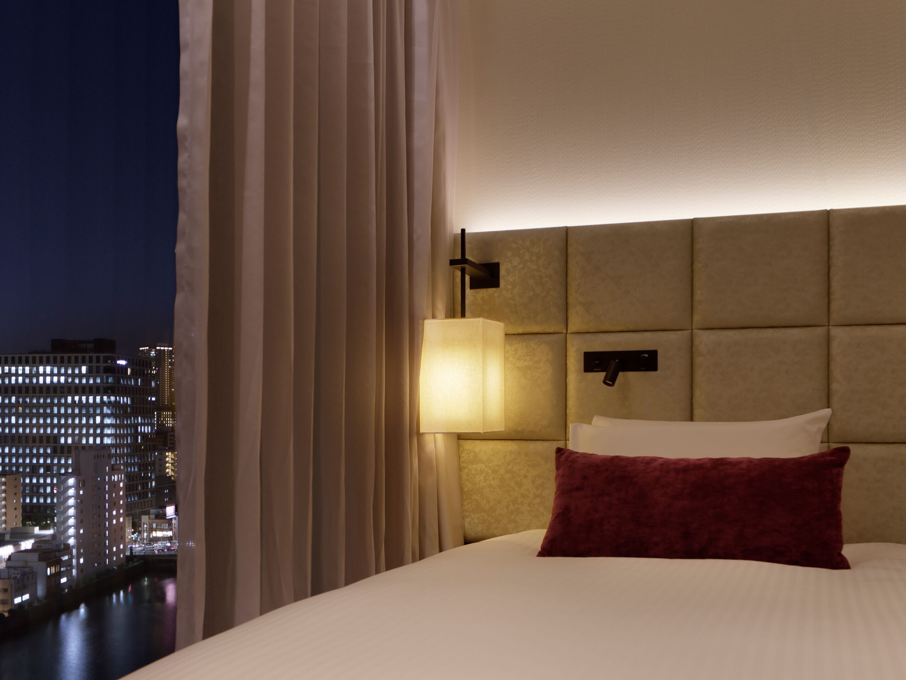 A hotel room with a large bed, a headboard with a light fixture, a view of a cityscape at night, and a curtain partially drawn.