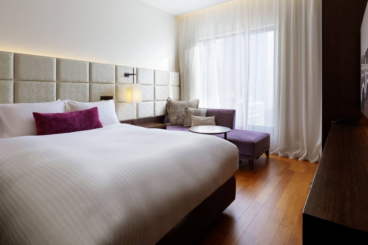 Une chambre d’hôtel avec un grand lit, une table de chevet, une petite table ronde, un canapé et une grande fenêtre avec des rideaux légers.