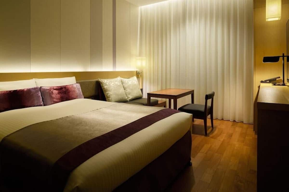 A hotel room with a bed, a desk, a chair, and a lamp.