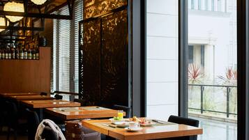 A restaurant with wooden tables and chairs, a bar area with bottles, and a view of buildings through large windows.