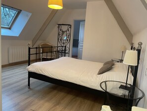 A bedroom with a bed, a glass table, a lamp, a radiator, a chair, and a window with a view of trees. - Château de Grèges (Greges)