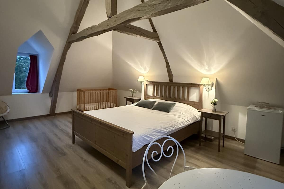 Un dormitorio con techo inclinado, vigas de madera, una cama, lámparas de noche, una mesita y una ventana con vistas.