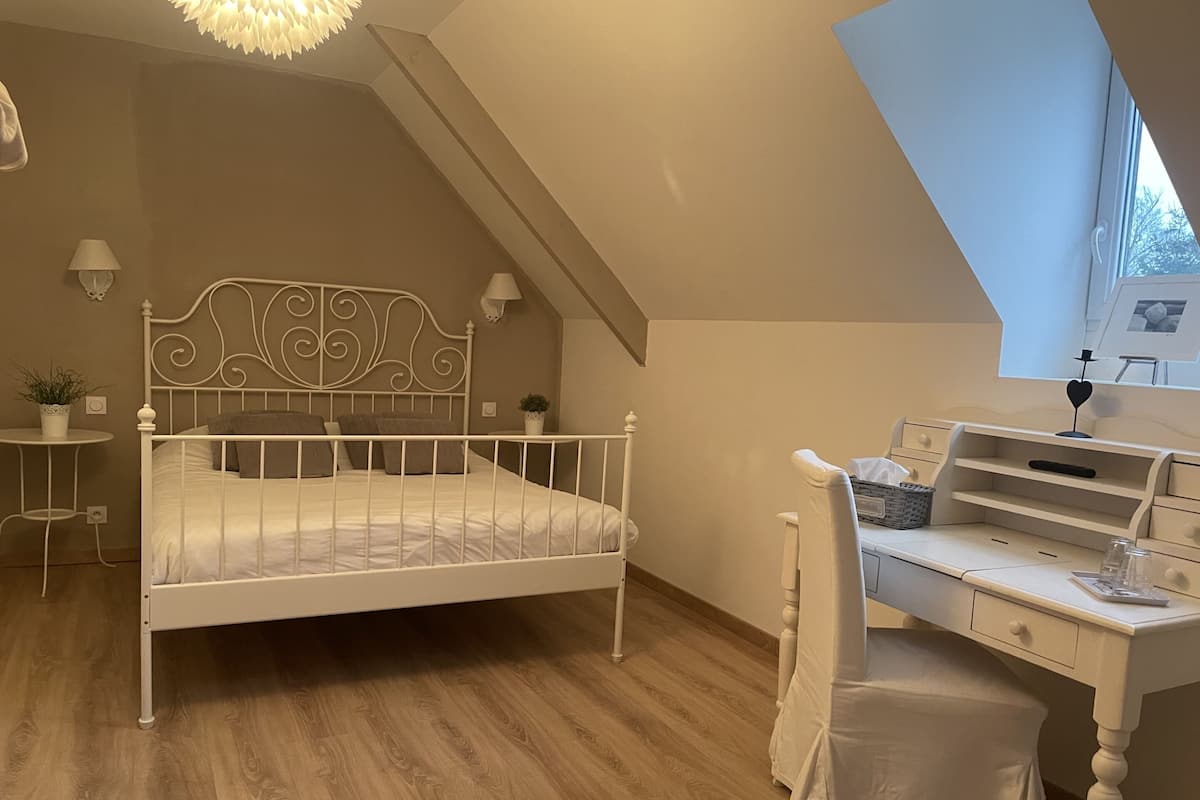 A bedroom with a white metal bed frame, a wooden desk with a chair, a small table with a lamp, and a potted plant.