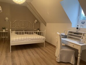 A bedroom with a white metal bed frame, a wooden desk with a chair, a small table with a lamp, and a potted plant. - Château de Grèges (Greges)