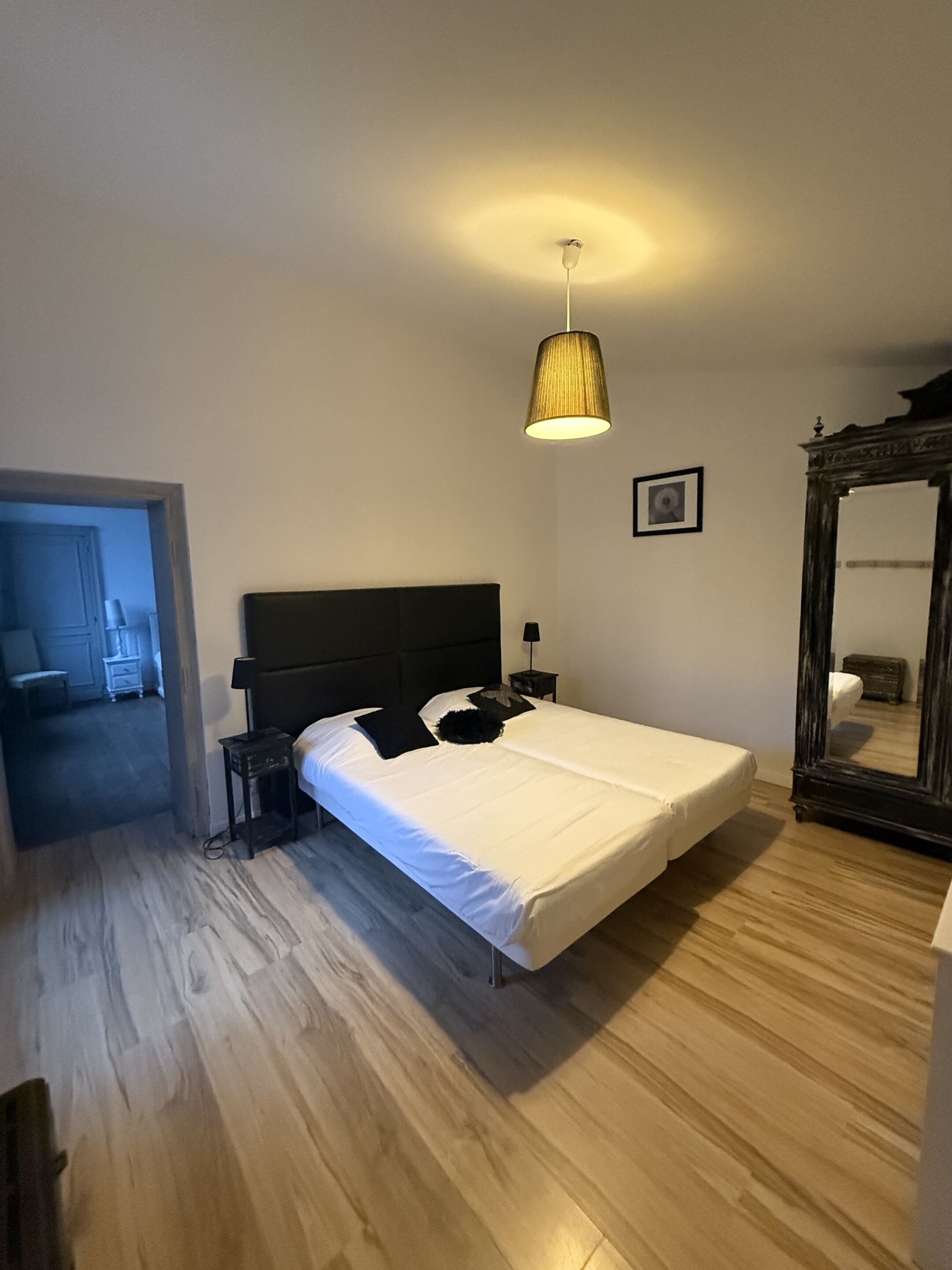 A bedroom with a bed, a bedside table, a lamp, a mirror, and a door leading to another room.