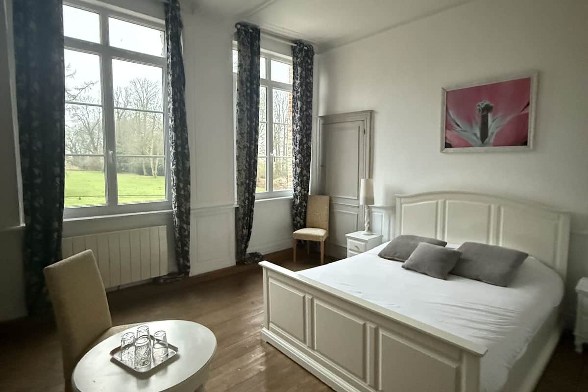 A bedroom with a large bed, a chair, a small table, a chandelier, a window with curtains, and a painting on the wall.