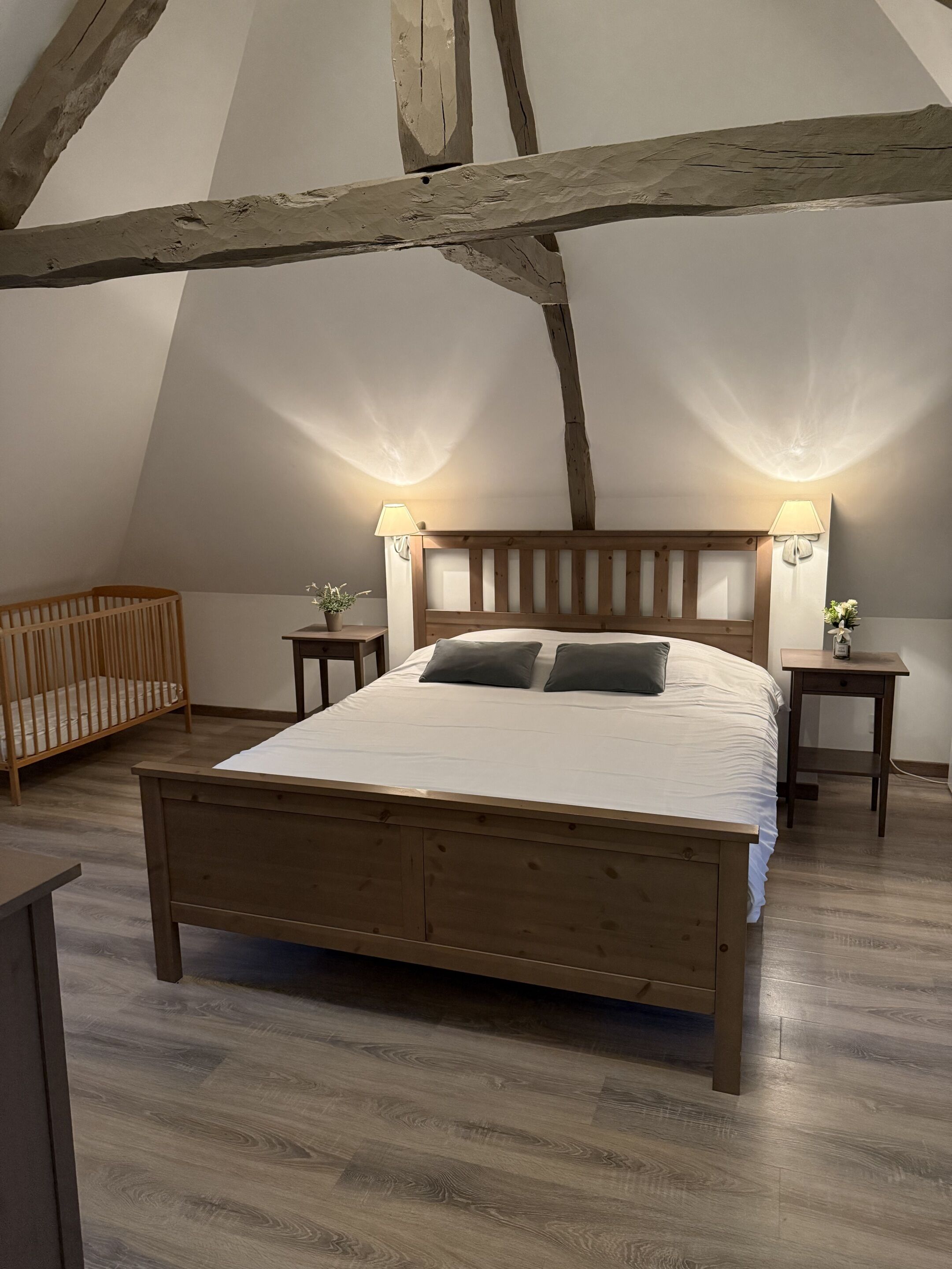 A bedroom with a wooden bed, two bedside tables, a nightstand, a crib, and a sloped ceiling with exposed beams.