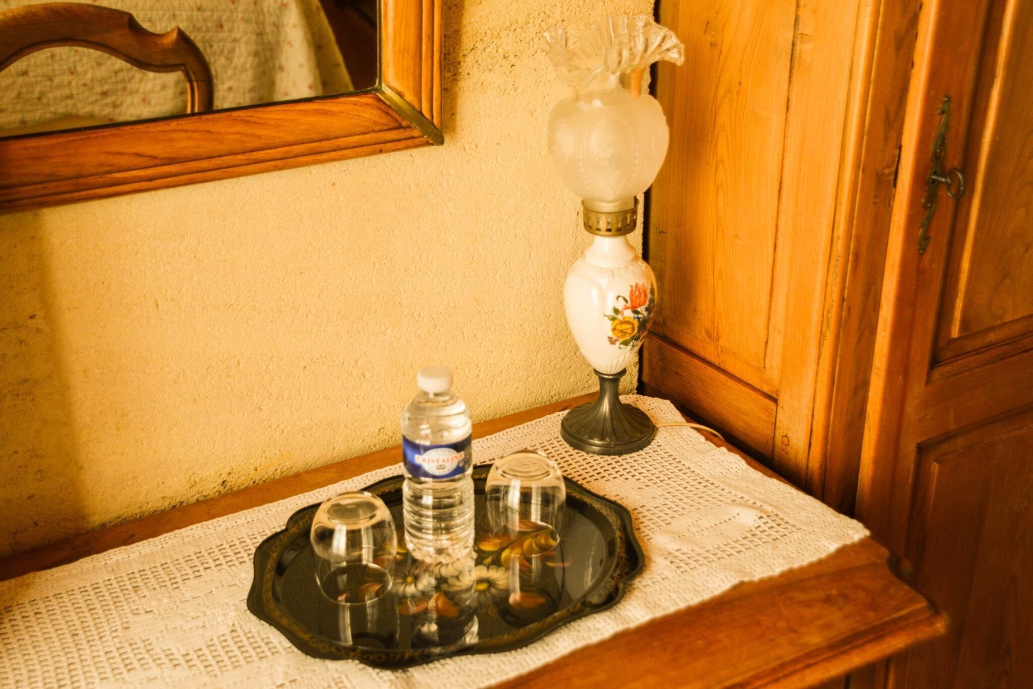 A wooden cabinet with a mirror above it, a table with a lace tablecloth, and a tray with a water bottle, salt shakers, and pepper shakers.