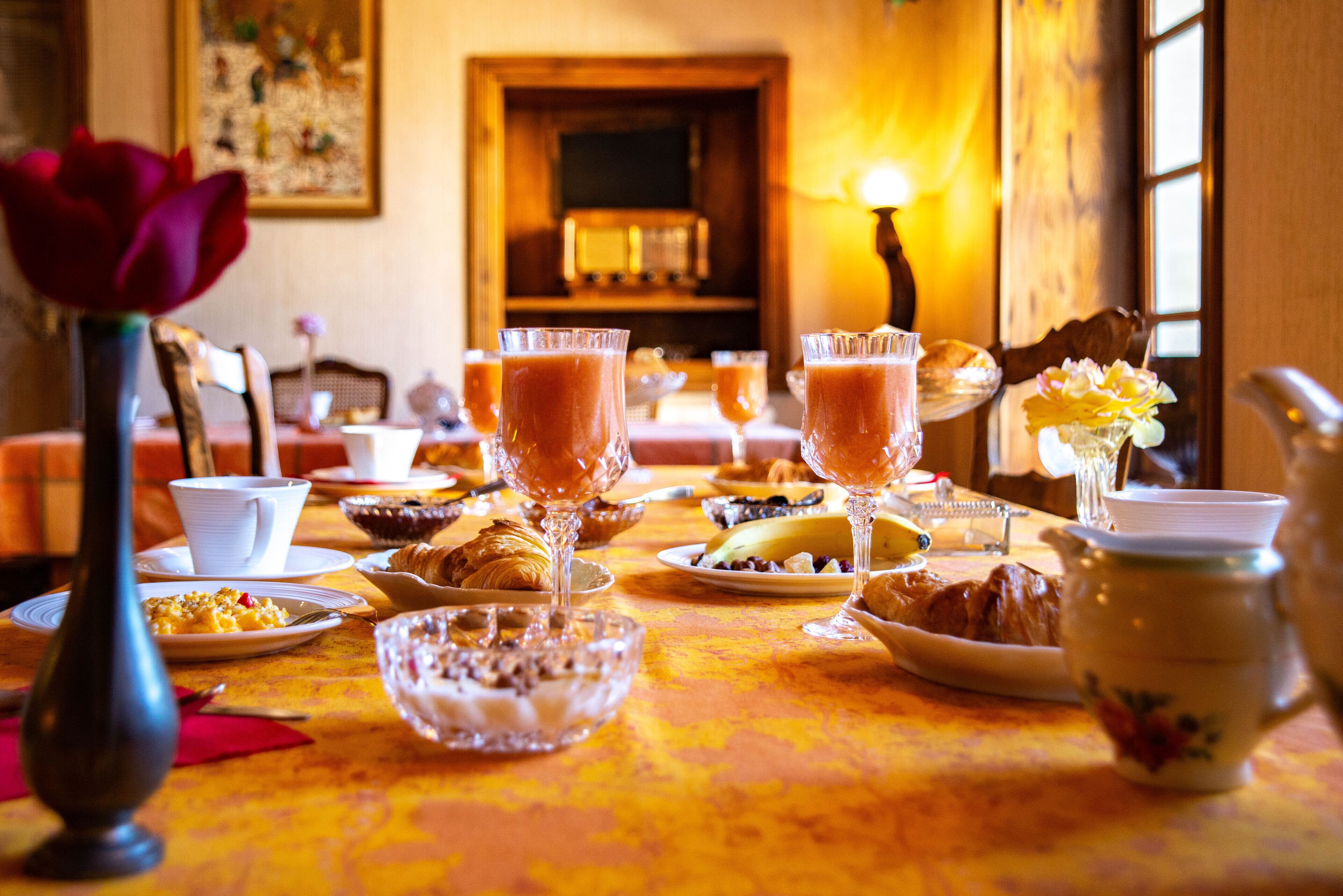 A breakfast setting with a table set for two, featuring croissants, fruit, and a vase with a flower.