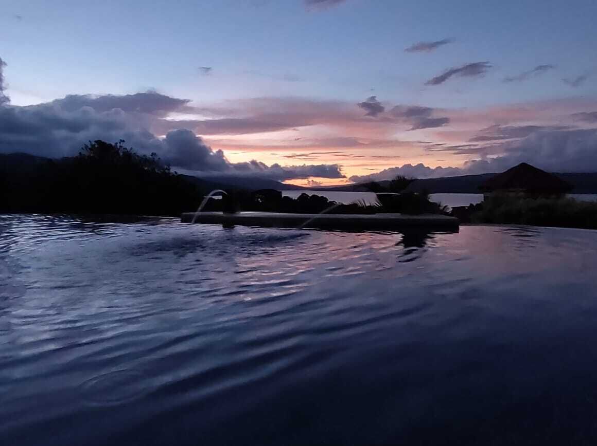 A serene pool area with a view of a sunset over a body of water, surrounded by vegetation and a distant mountain range.