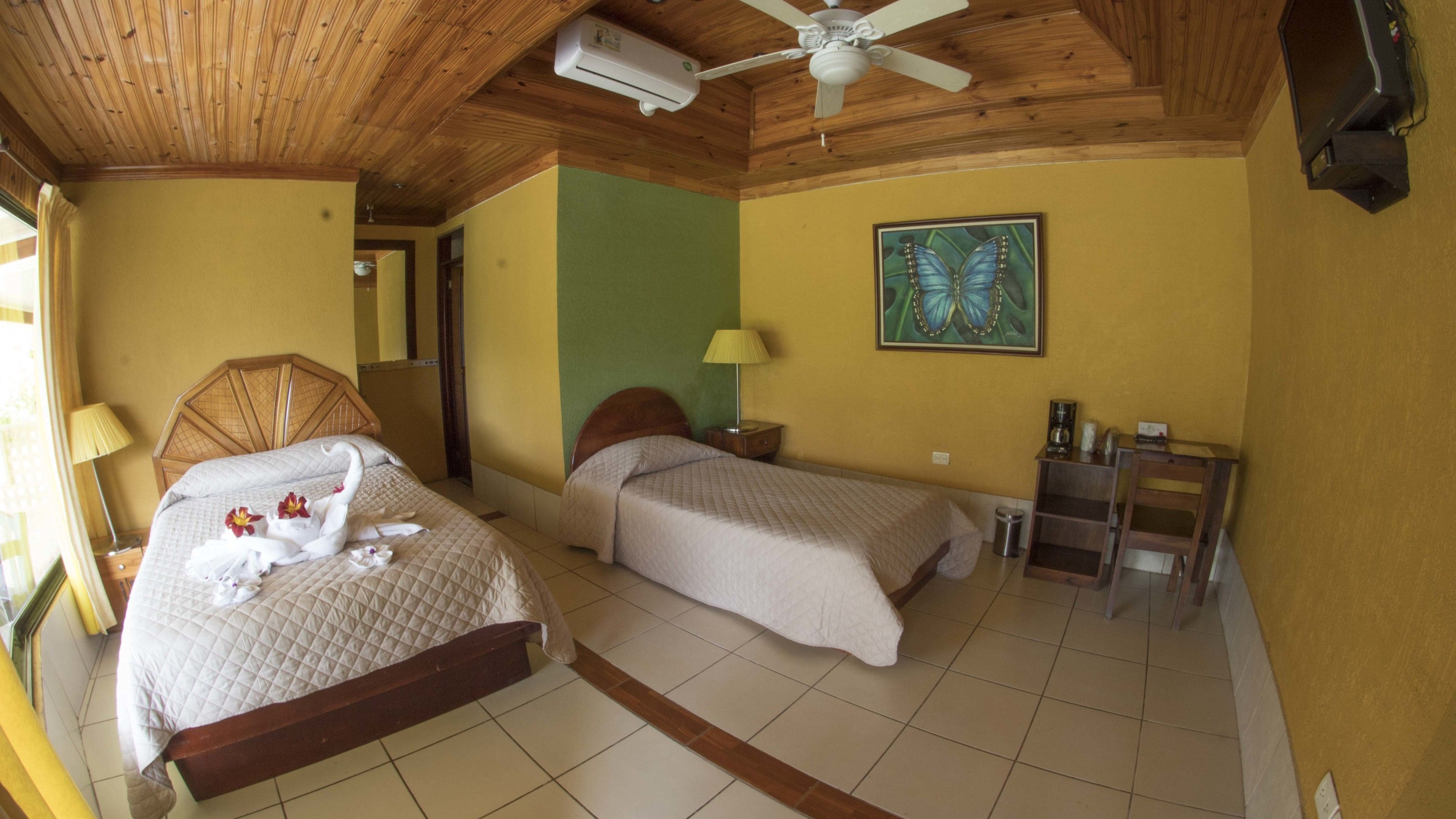 Habitación de hotel con dos camas, ventilador de techo, un cuadro y una pequeña mesa con lámpara.